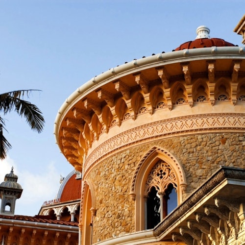 Ornate stone building with domed roofs, arched windows, and intricate detailing, framed by palm tree fronds against a clear sky.