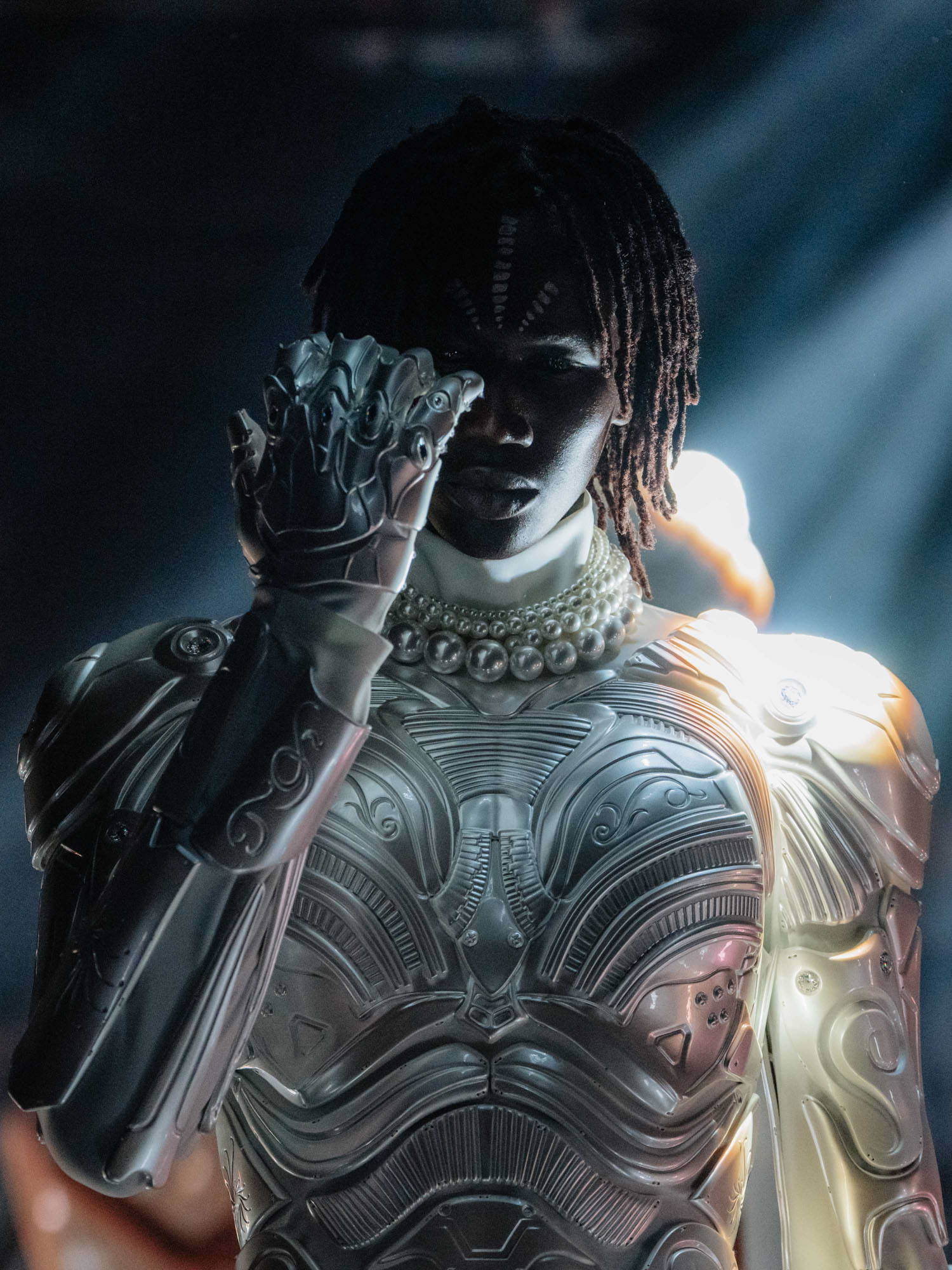 A person wearing an intricate, light-reflecting metallic suit and a pearl necklace poses dramatically under moody lighting with one hand partially covering their face.