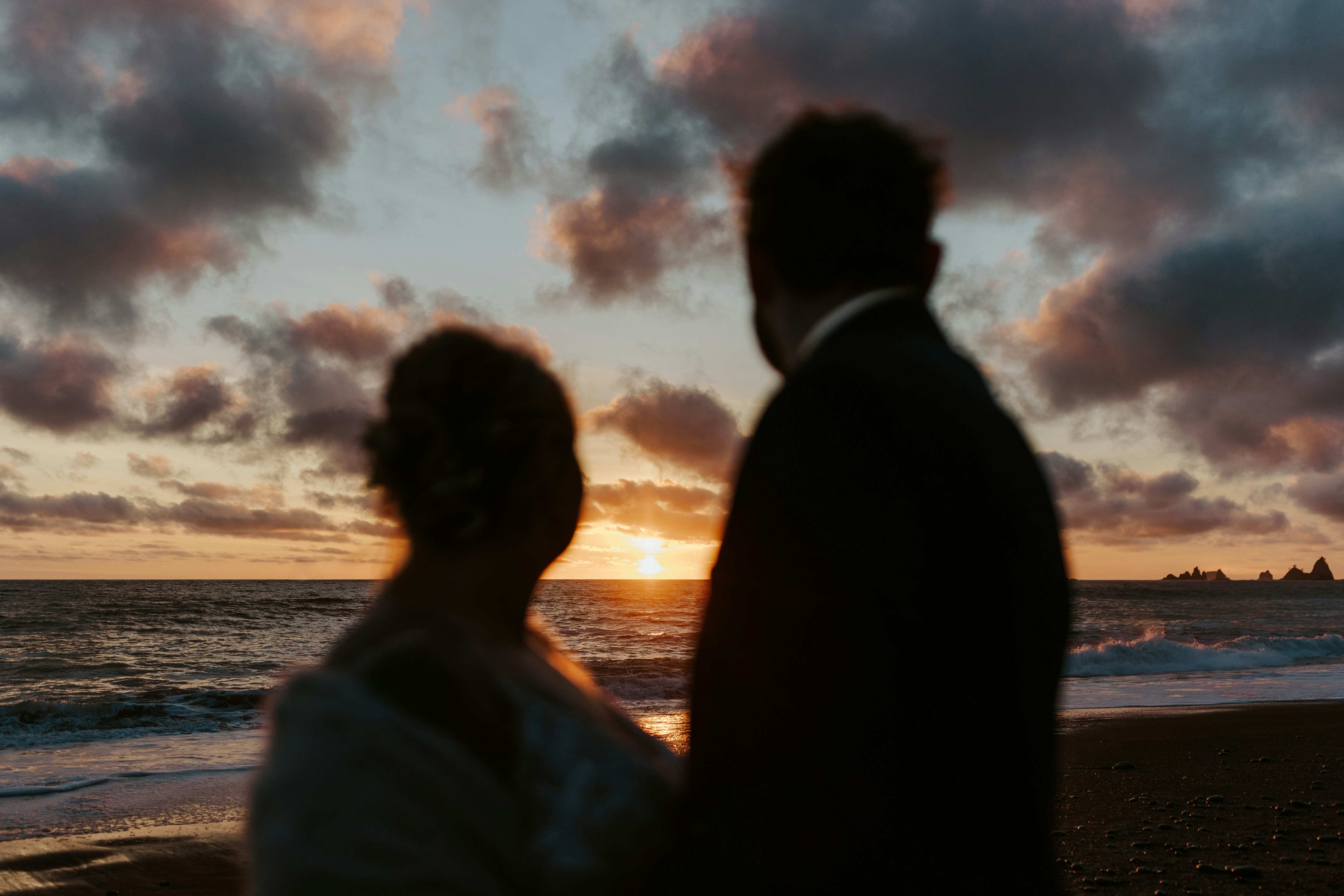 a bride and groom standing on the beach at sunset