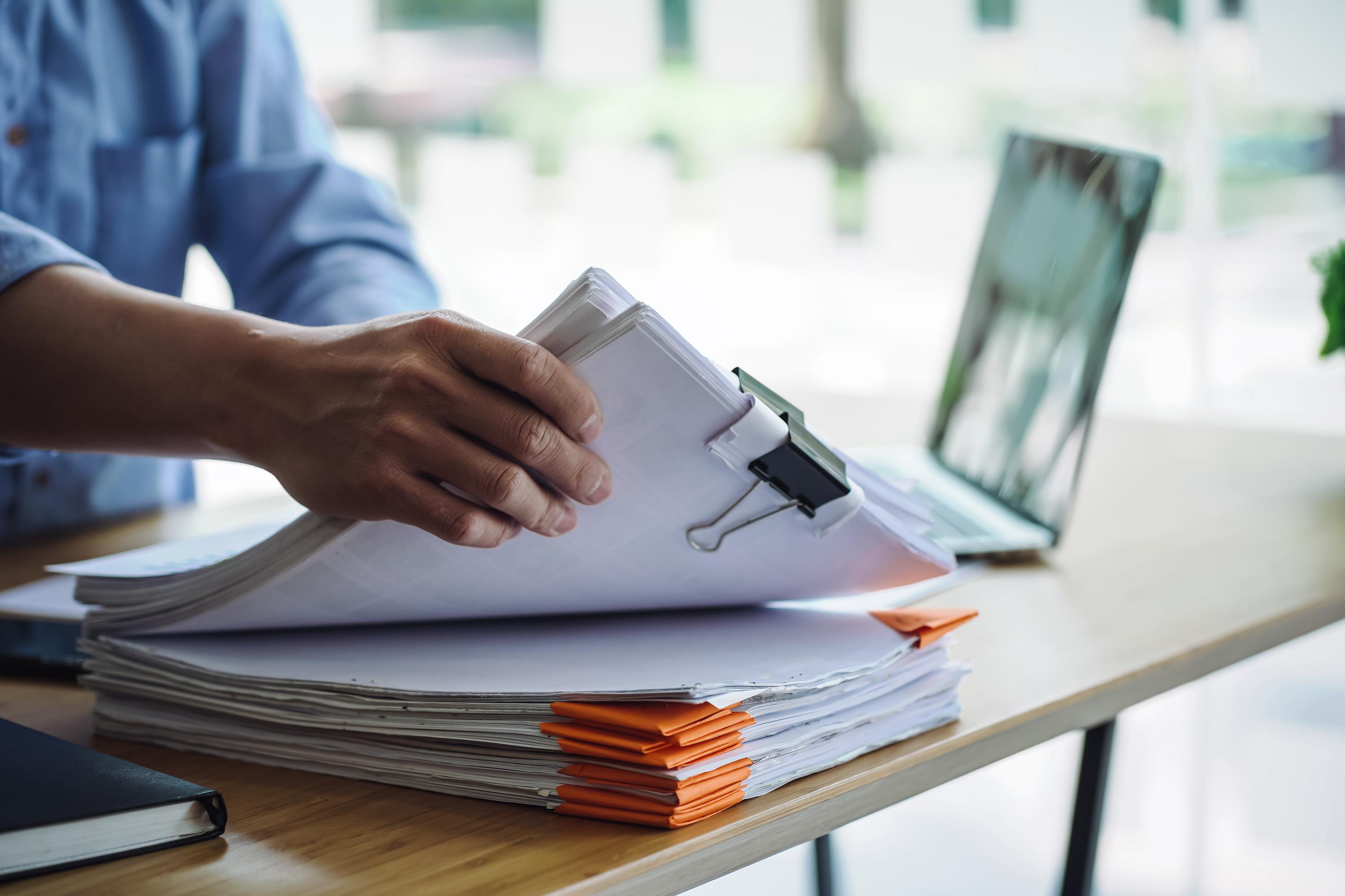 Close-up of a person reviewing a large stack of paper documents secured with a binder clip and marked with orange tabs on a desk, with an open laptop in the background.