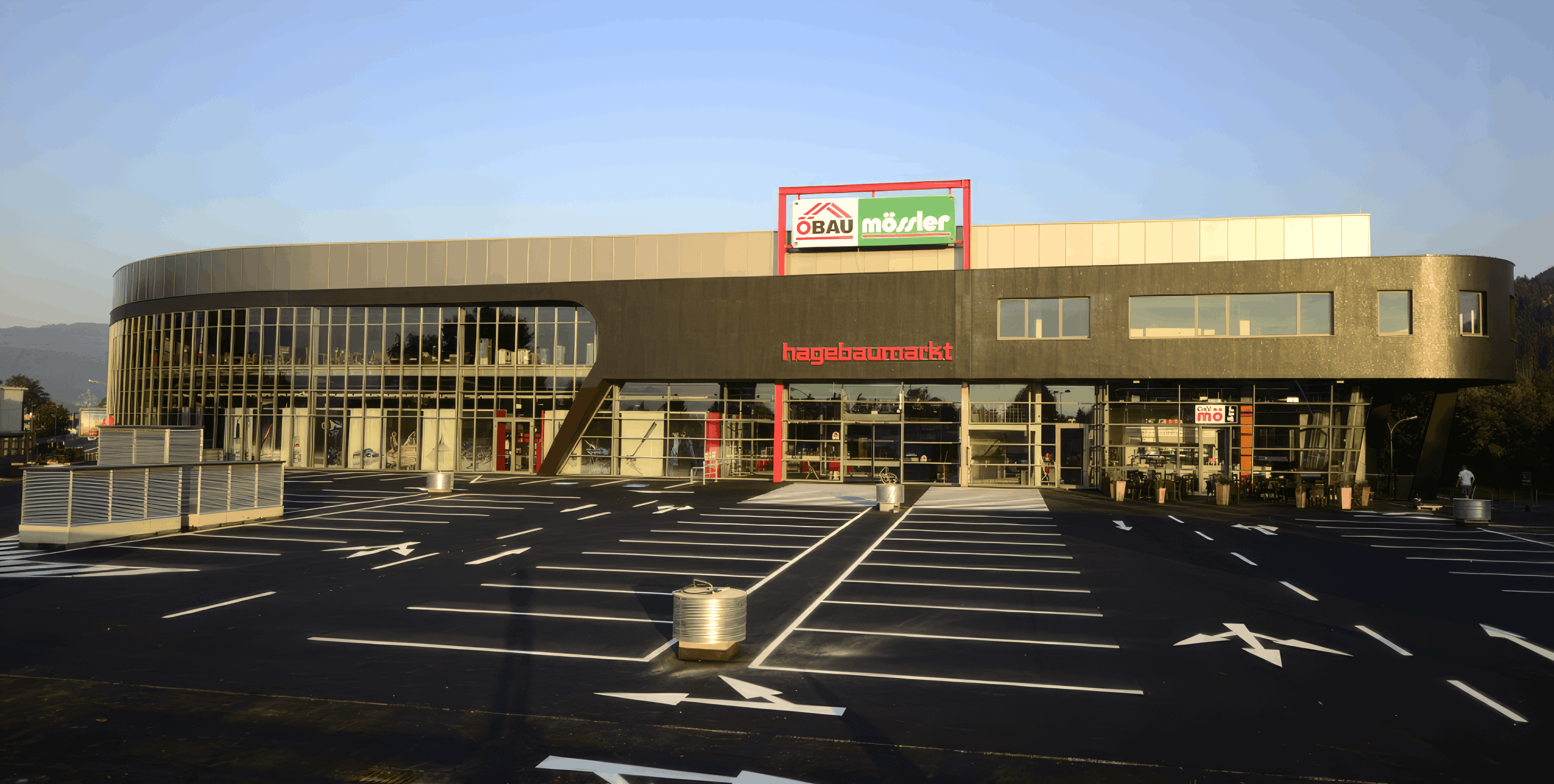 MW Interior - Hagebaumarkt Außenansicht mit geschwungener dunkler Fassade, Glasfront und großem Parkplatz im Abendlicht" Beschreibung: Moderner Hagebaumarkt-Standort mit markanter Rundform-Fassade und weitläufigem Kundenparkplatz – ein Referenzstandort im Bau- und Heimwerkhandel, für den Ladeneinrichtung und Möblierungslösungen realisiert wurden.