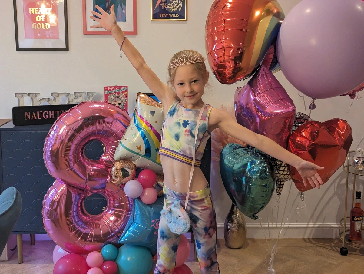 Girl posing with large pink and purple number 8 balloon stack and heart balloons indoors