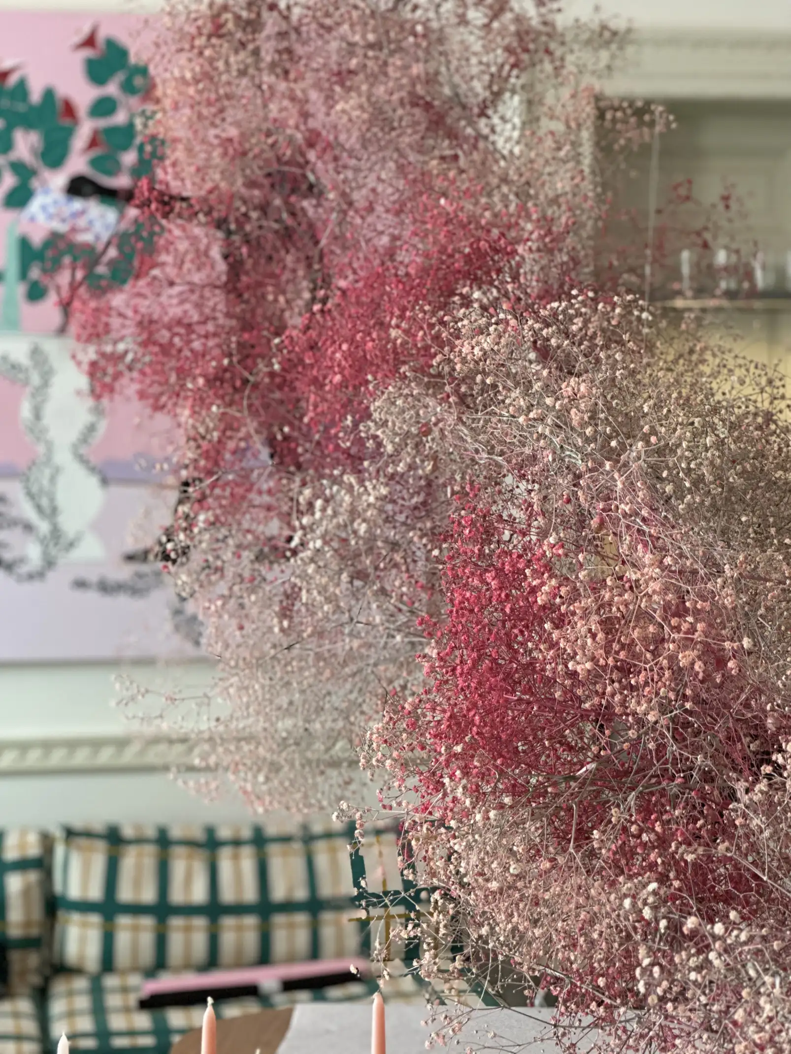 Instalación floral suspendida con ramilletes de flores en tonos rosa y blanco, flotando sobre una mesa de madera cubierta con papel blanco, en una sala luminosa con molduras clásicas y cortinas de fondo.