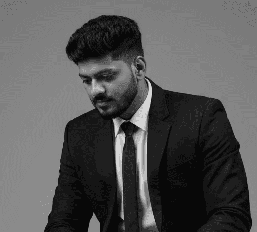Studio portrait of a young Afro-descendant man warmly smiling at the camera. He has a short beard, upward-styled hair, and is wearing a dark blue T-shirt against a neutral gray background.