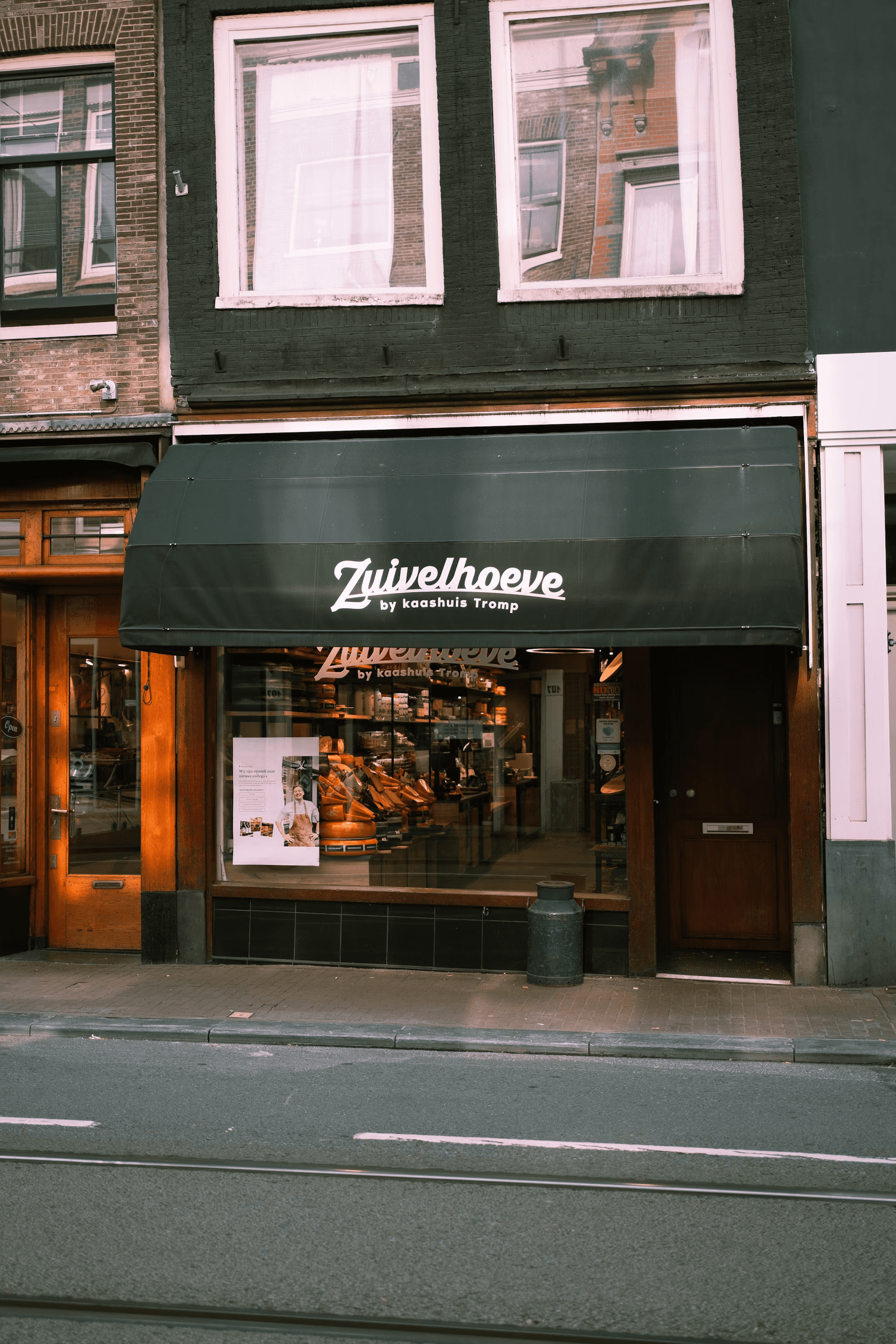 A quaint storefront with a black awning displaying the name "Zuivelhoeve," showcasing an inviting interior filled with shelves of products and a warm, welcoming atmosphere; ideal for those exploring local markets.