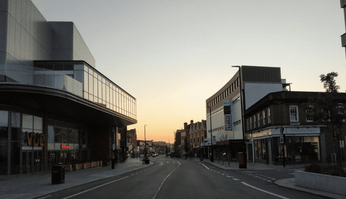 A smiling man with curly hair is sitting at a desk, looking at the camera, with a laptop and a lamp in the background.
