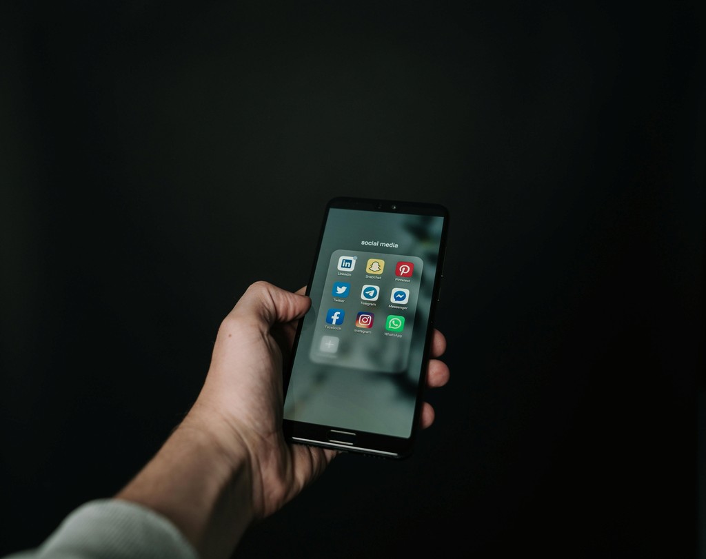 The image displays a hand holding a smartphone against a dark background, with the screen showing a folder labeled "social media" containing various app icons, including LinkedIn, Twitter, Pinterest, Facebook, Instagram, WhatsApp, Messenger, and YouTube. The minimalist and focused composition emphasizes the prevalence and importance of social media in modern communication.