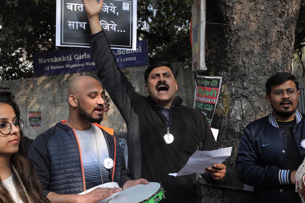 Smiling young woman with long hair standing against a dark green background, holding a finger to her chin.