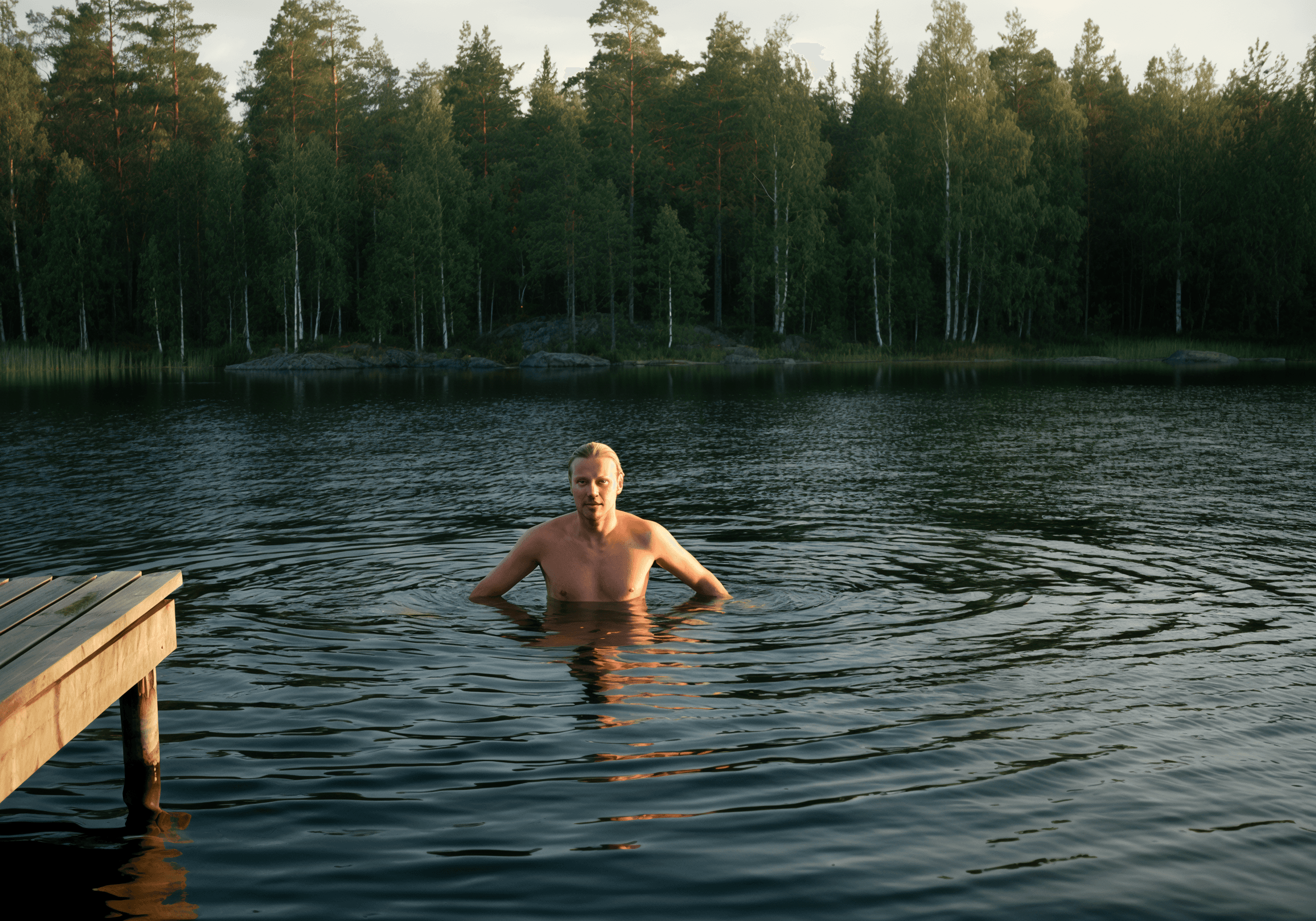 Man swimming alone in a calm forest lake, surrounded by trees at sunset.