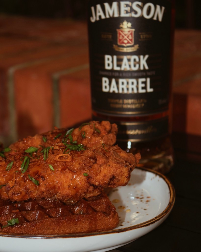A plate featuring two crispy fried chicken pieces on top of a waffle, garnished with green onions, accompanied by a bottle of Jameson Black Barrel whiskey in the background.