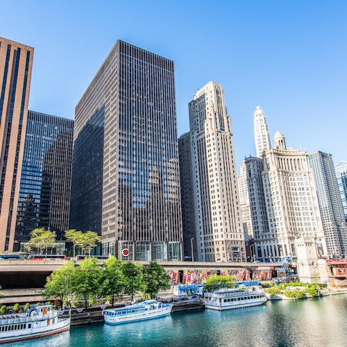 Tall buildings along a river, with boats docked beside lush green trees, under a clear blue sky.