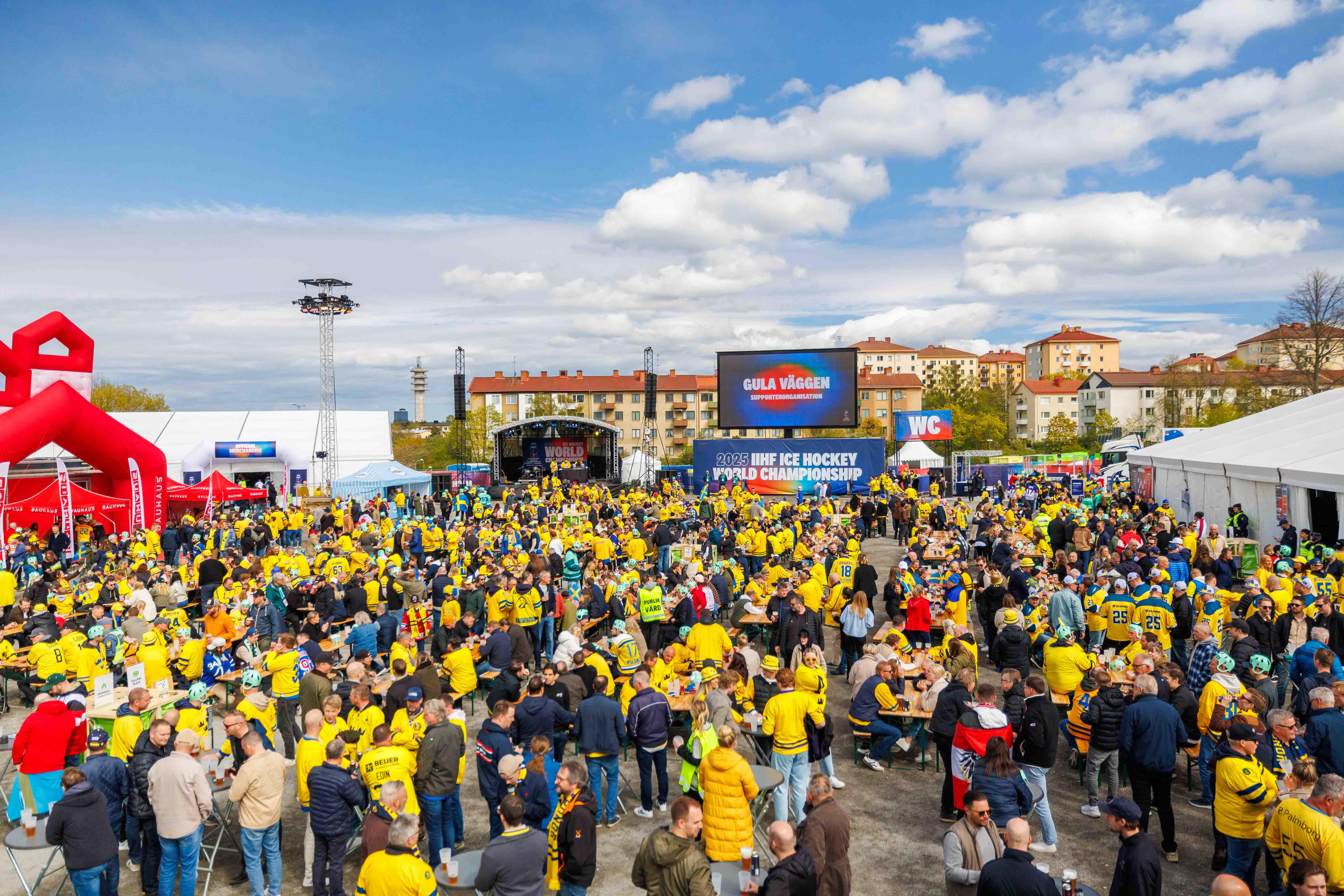 Crowds dressed in yellow gather at the fan zone during the IIHF 2025 Ice Hockey World Championship with LED screens and stage setup.