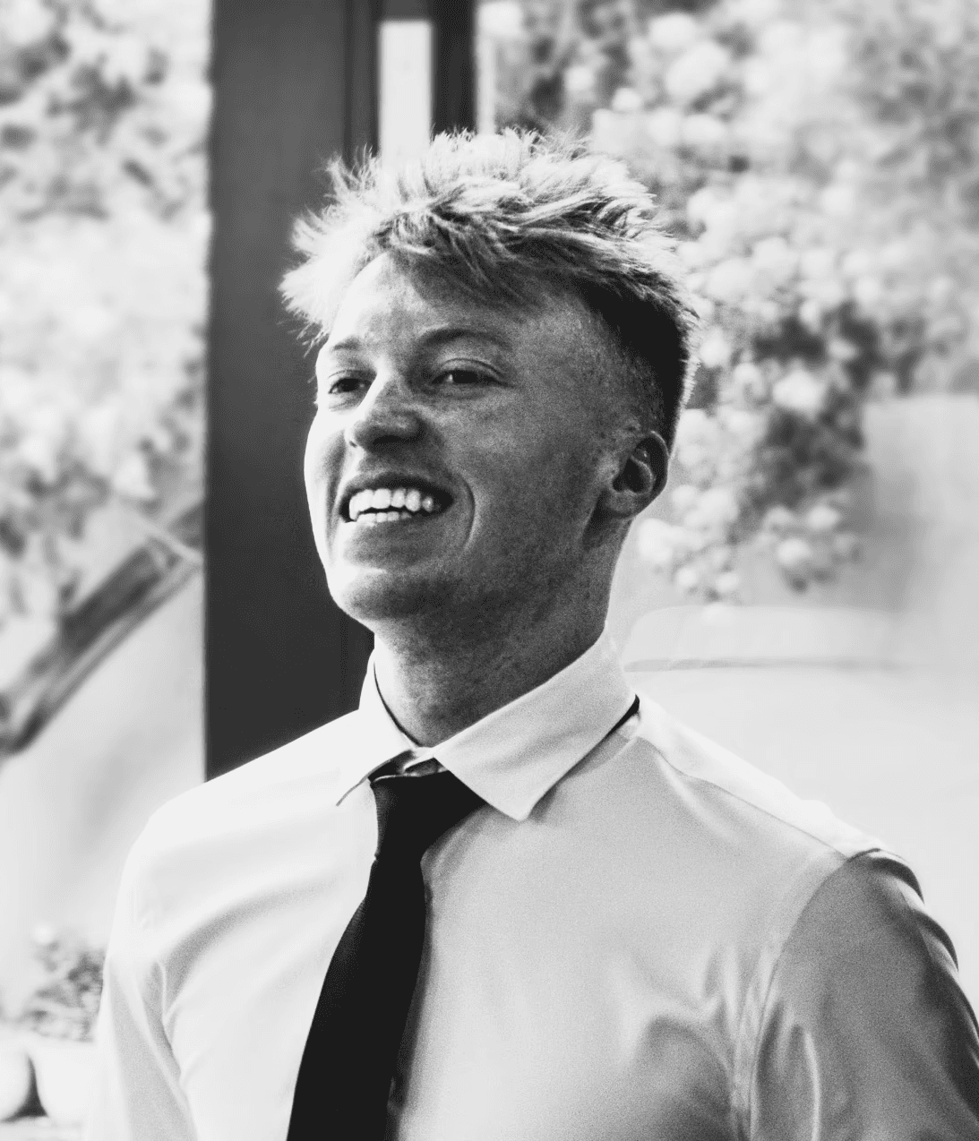 A smiling person with blonde, wavy hair, wearing a white shirt and black tie, stands in a bright environment.