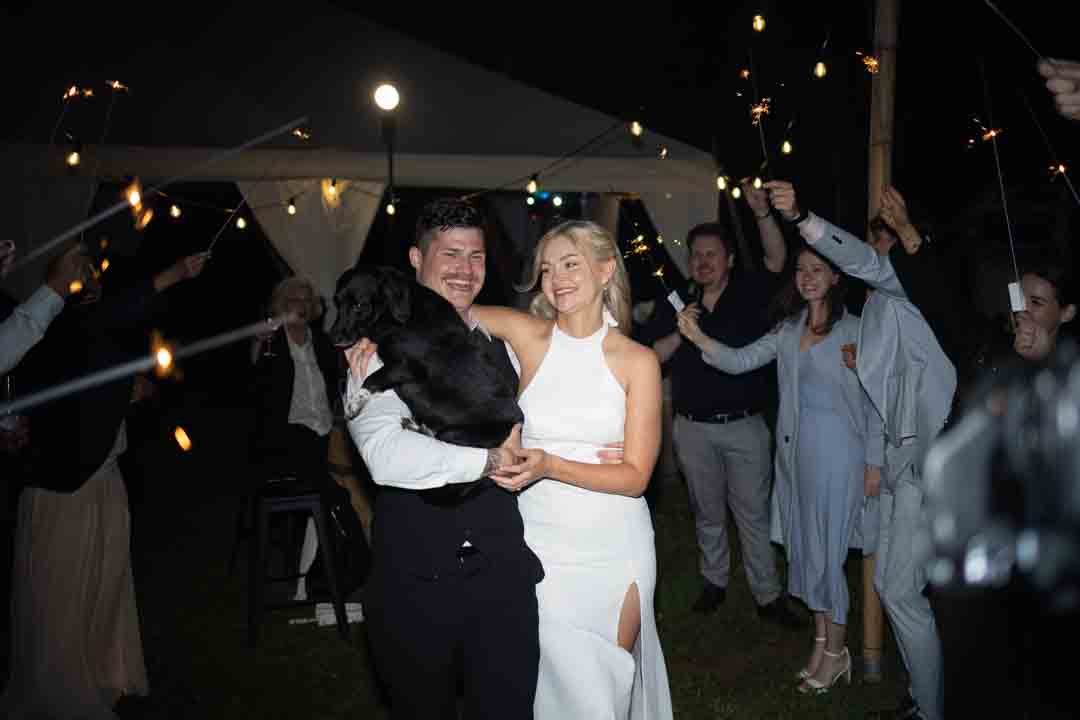 Groom and bride holding black dog walking through guard of honour with sparklers