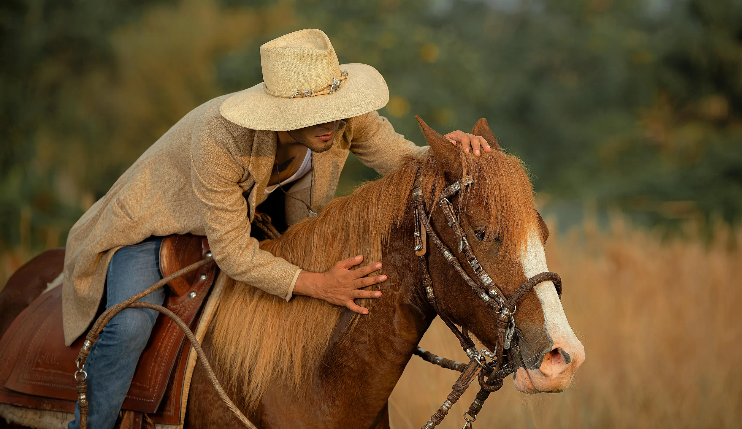 Person riding a chestnut horse outdoors during golden hour, wearing a straw hat and beige jacket