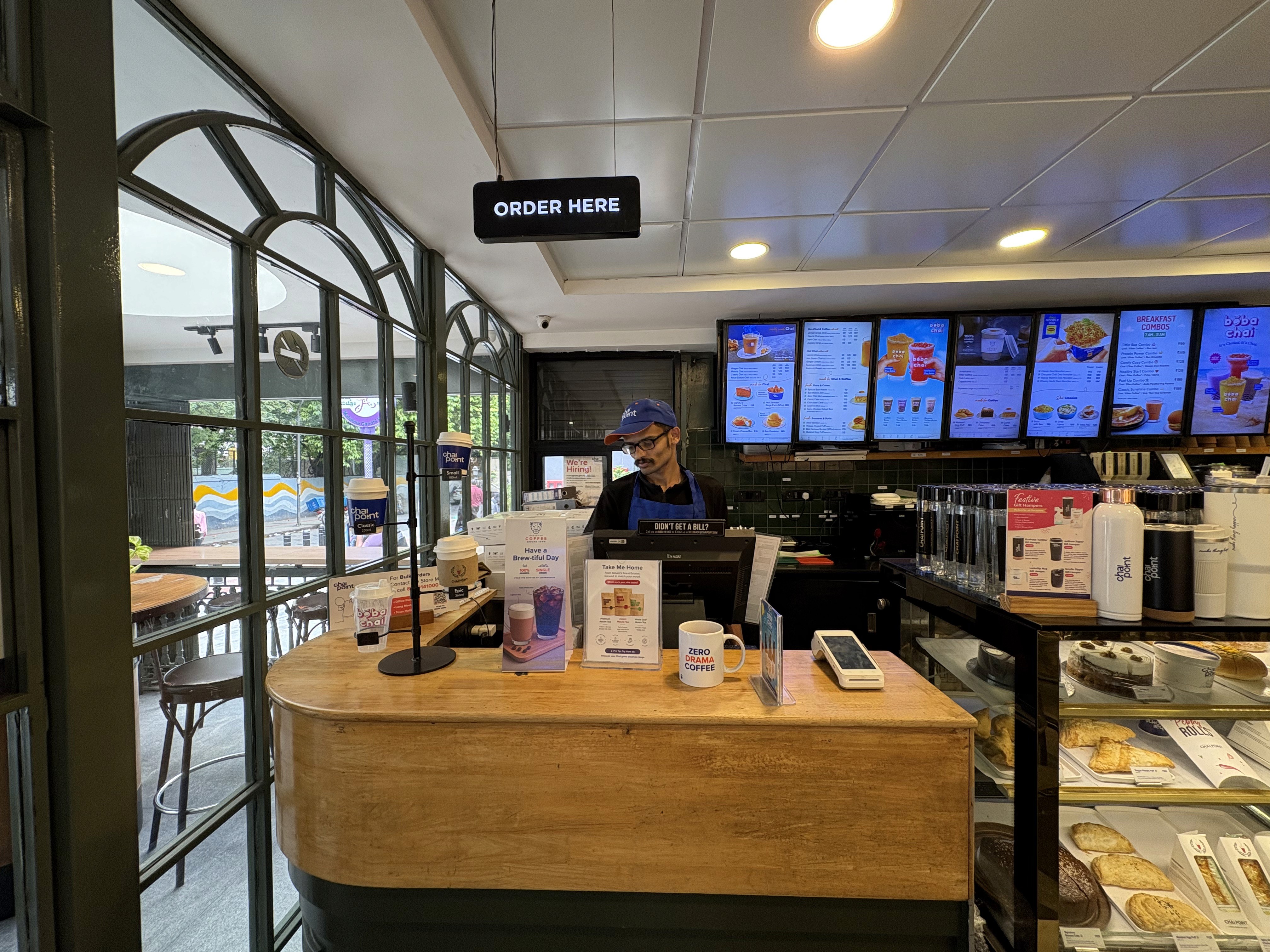 A Chai Point staff member stands behind the counter in uniform, surrounded by takeaway cups, promotional displays. Digital menu boards show drink and snack options, in Bangalore.