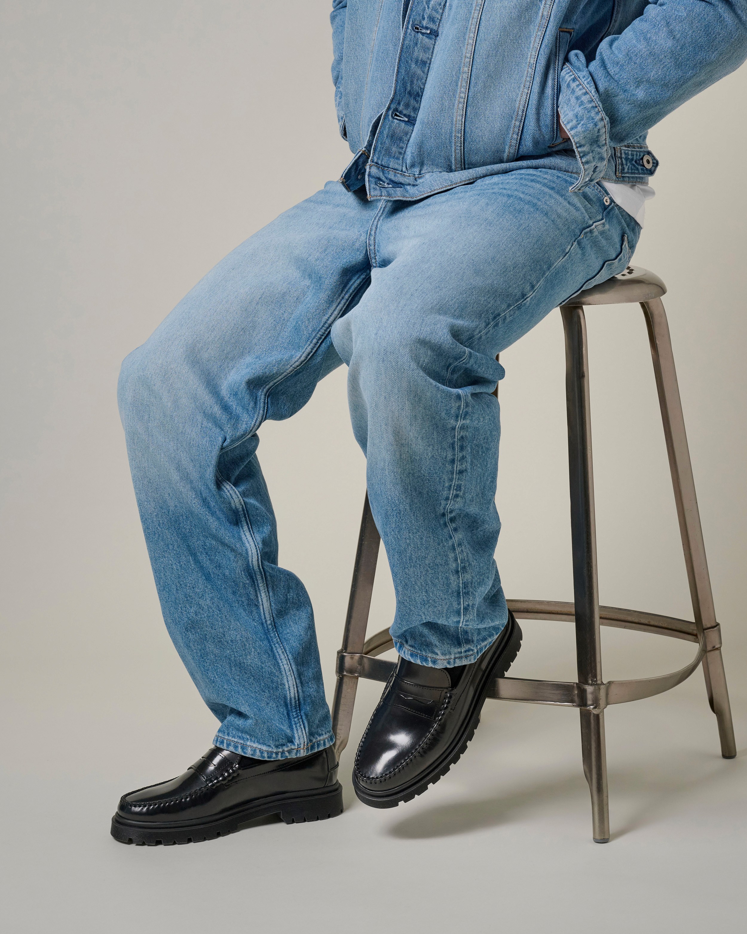 Le bas d'une silhouette assise sur un tabouret, portant du jean délavé et une paire de mocassins penny noirs vernis