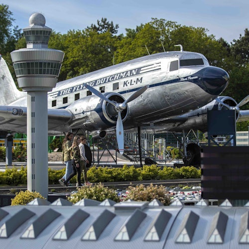 Two people walking near a silver plane labeled "The Flying Dutchman" with a control tower and green foliage in the background.
