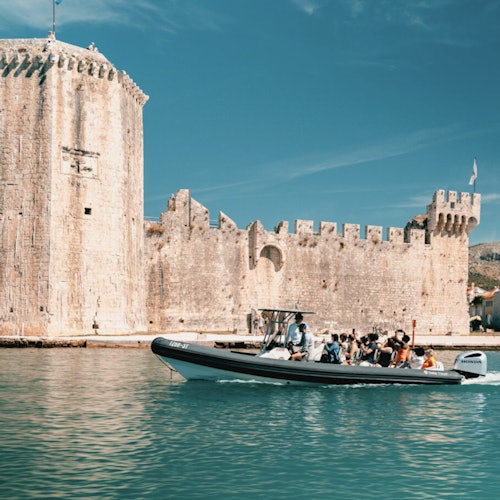 En liten båt med folk passerer en historisk steinborg på en solfylt dag, med klart blått vann og himmel i bakgrunnen.