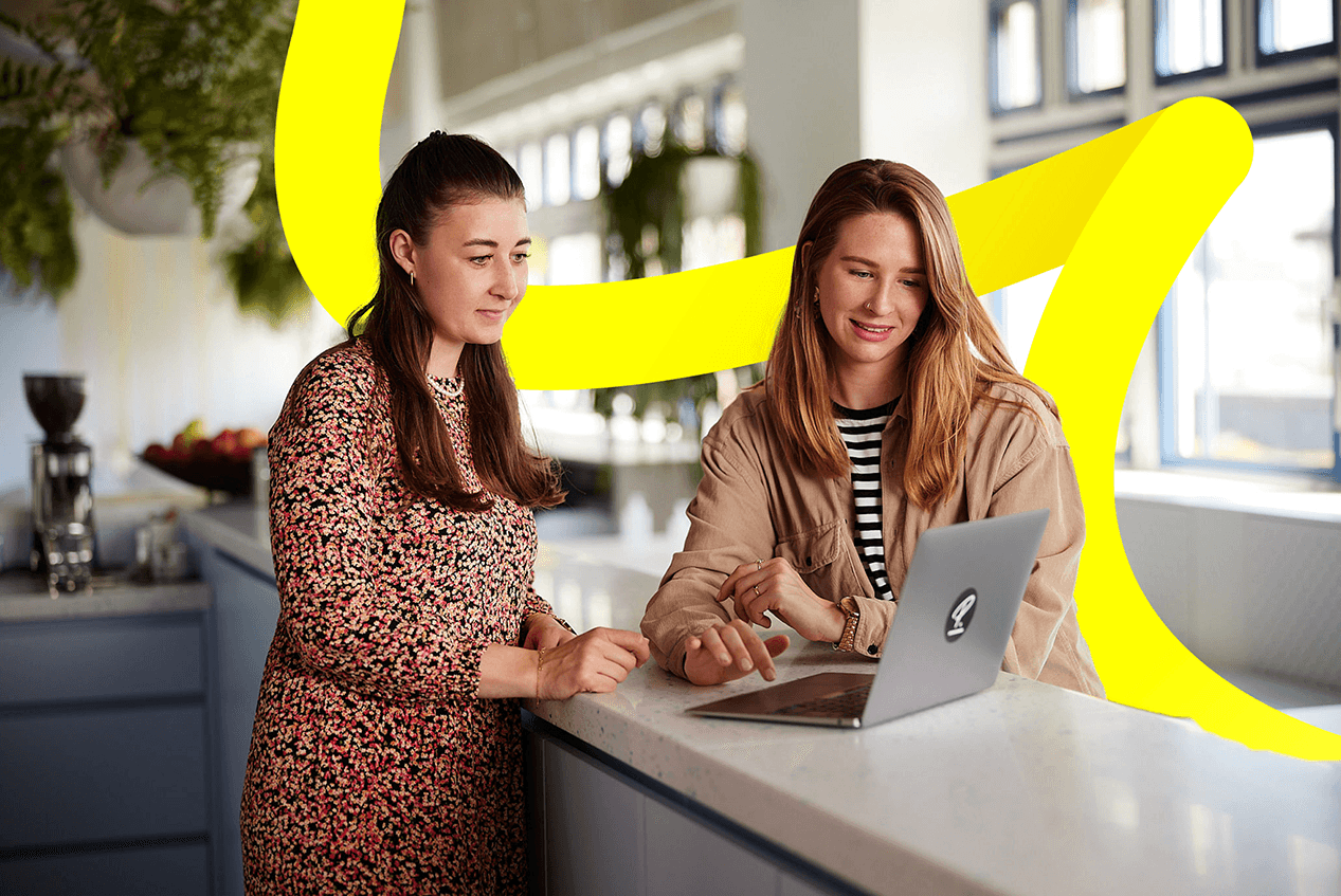 Twee vrouwen glimlachen en werken samen achter een laptop in een lichte, moderne keuken.