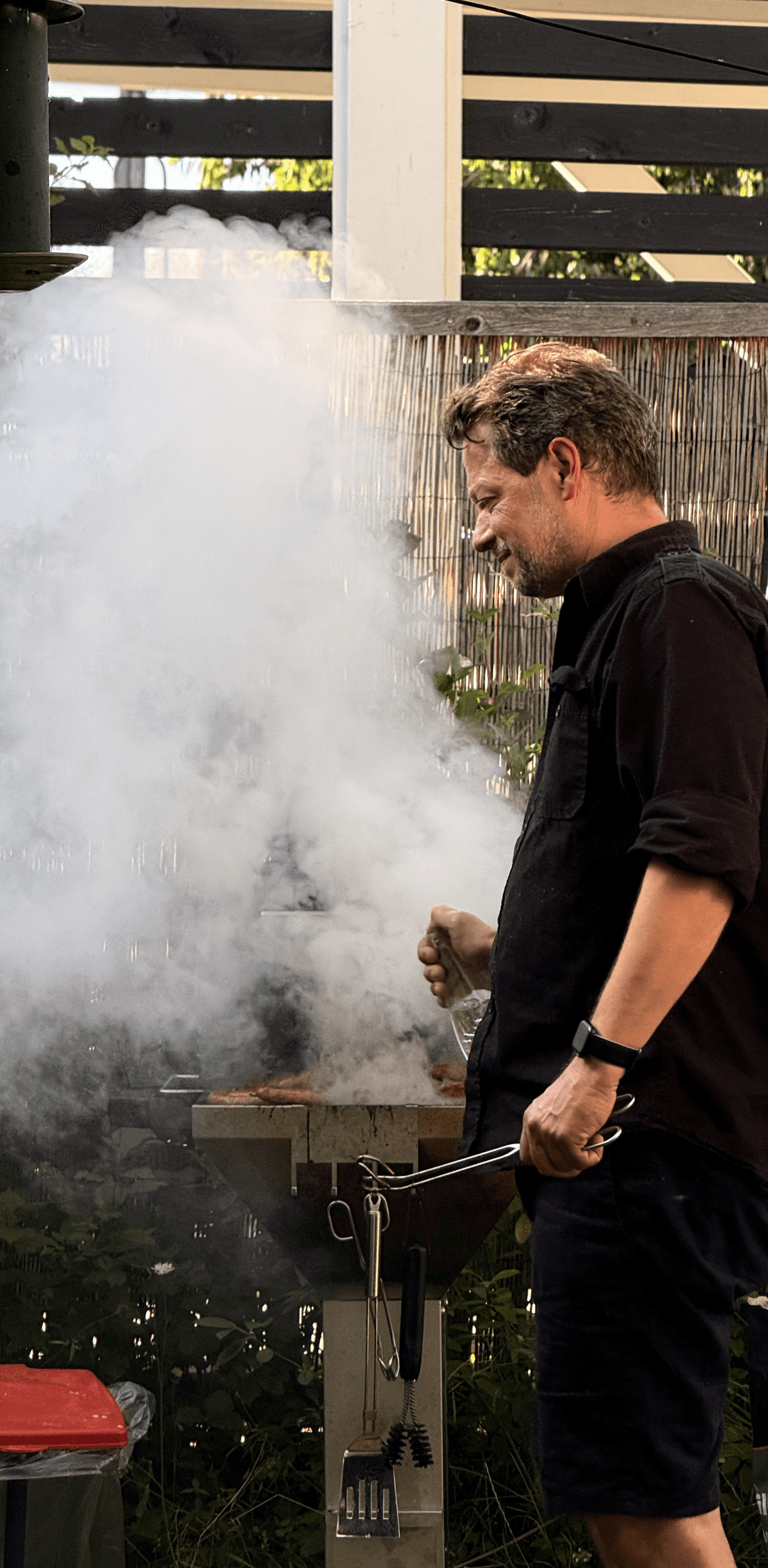 Mann in schwarzer Kleidung steht an einem rauchenden Grill im Garten
