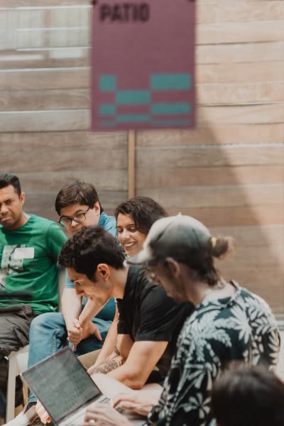 A group of people are sitting and smiling in a casual outdoor setting under a patio sign. They appear relaxed and engaged, with one using a laptop.