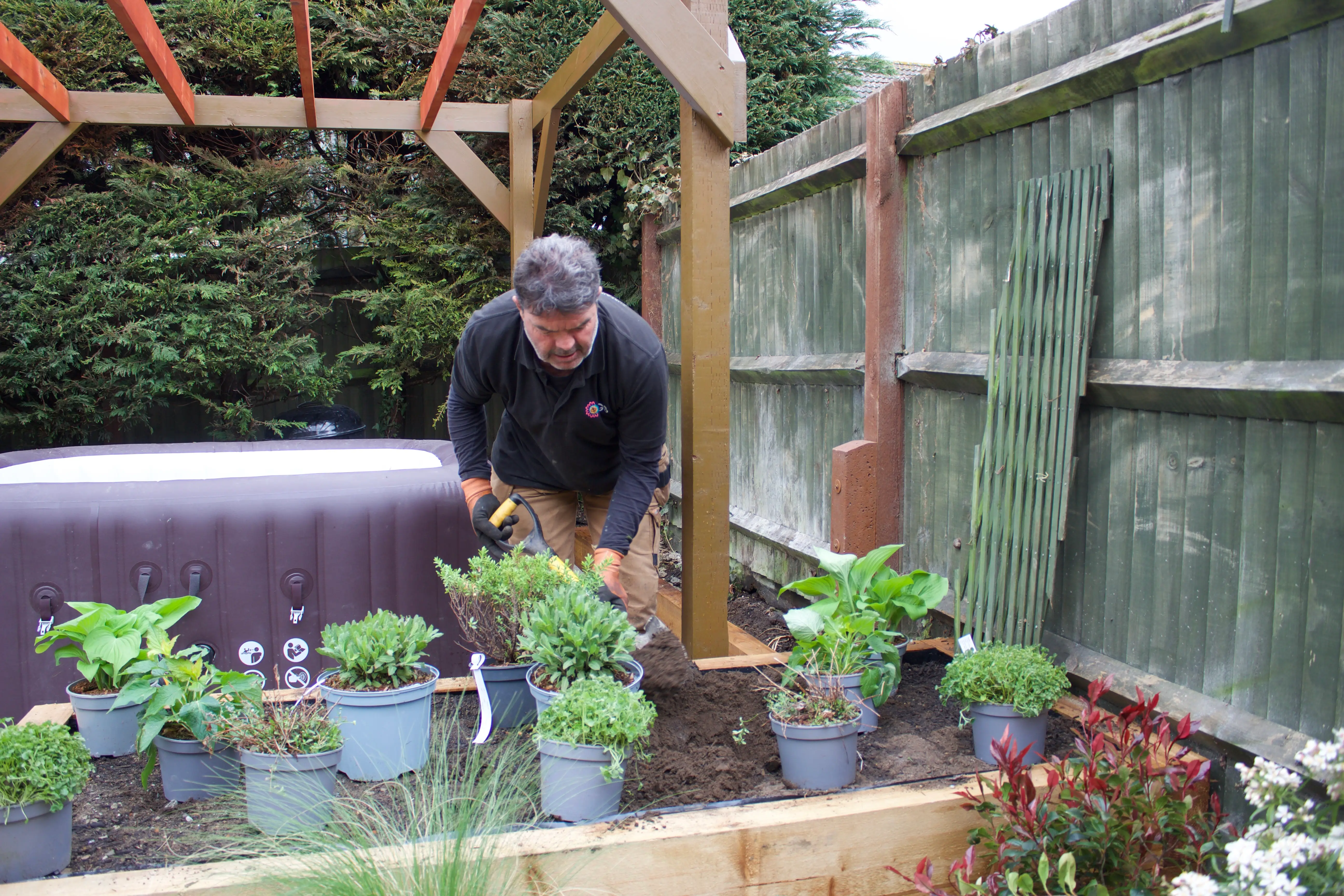 A person tending to potted plants in a garden, with a wooden structure and fence in the background.