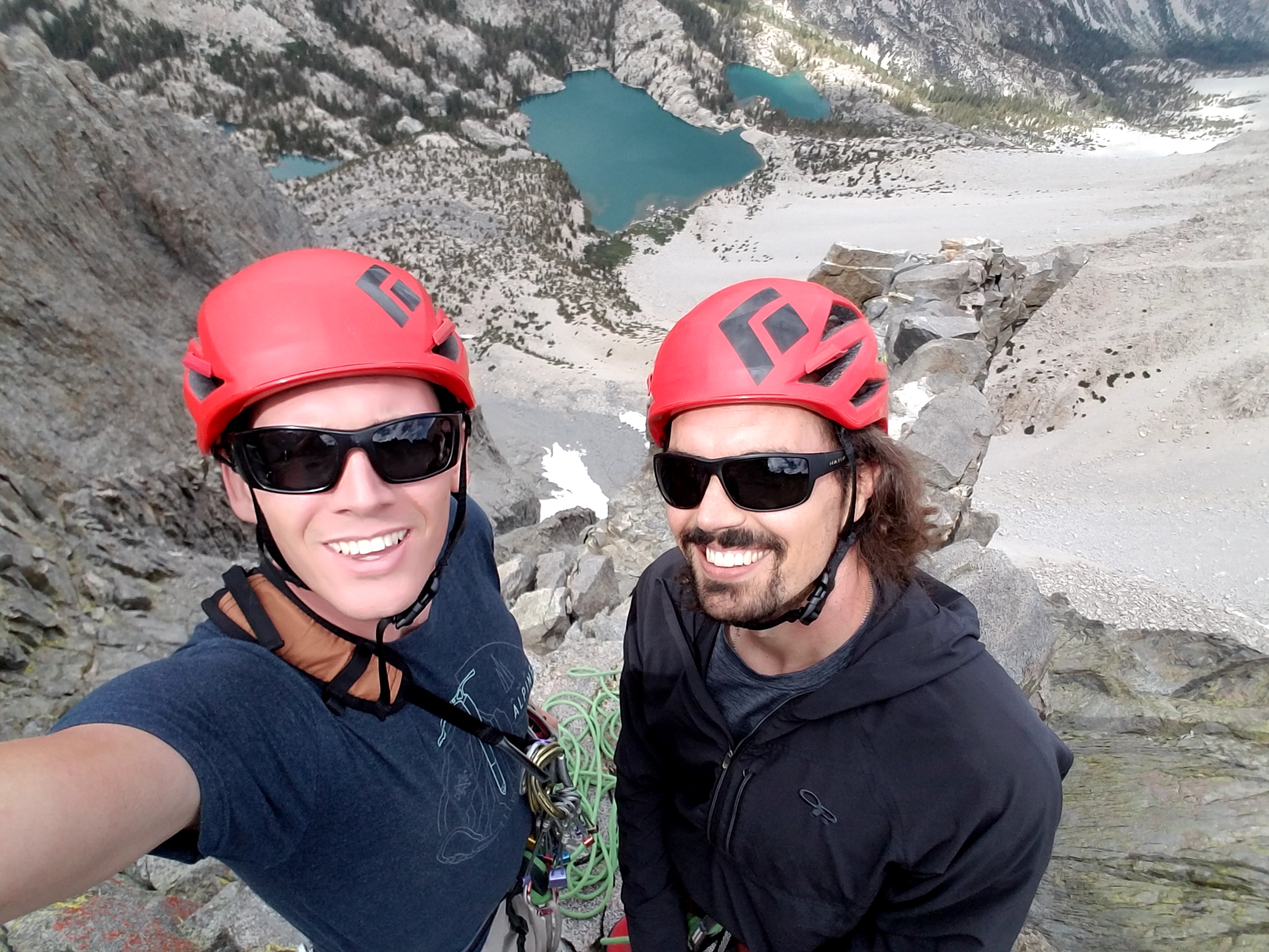 Guide and climber smiling with landscape opening up below with alpine lakes and crags