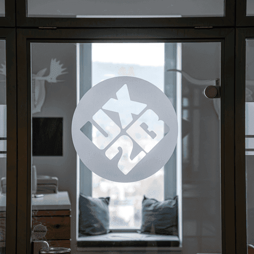 Glaswand mit rundem Aufkleber, der das UX2B-Logo in weiß zeigt, im Hintergrund ein Fenster mit Sitzbank und Kissen sowie zwei Tier-Figuren an der Wand