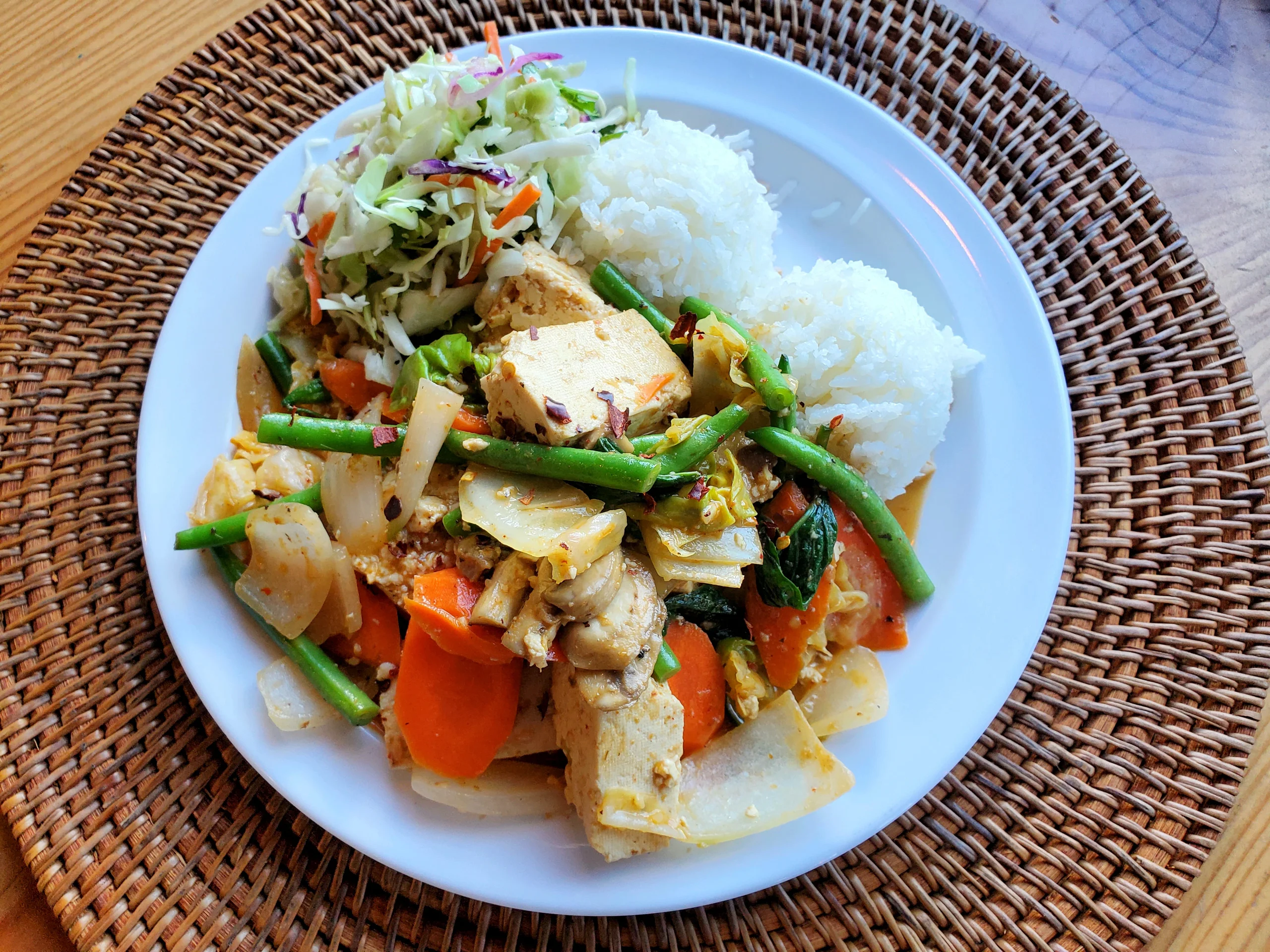 A white plate featuring a colorful stir-fry of tofu, green beans, and carrots in a red curry sauce, served with two scoops of white rice and a side of Asian cabbage slaw.
