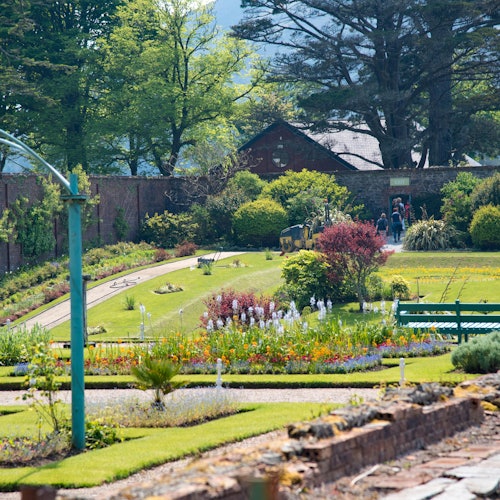 Добре поддържана градина с цветни цветни лехи, пътеки, пейки и дървета, в която се виждат хора на заден план близо до сграда.