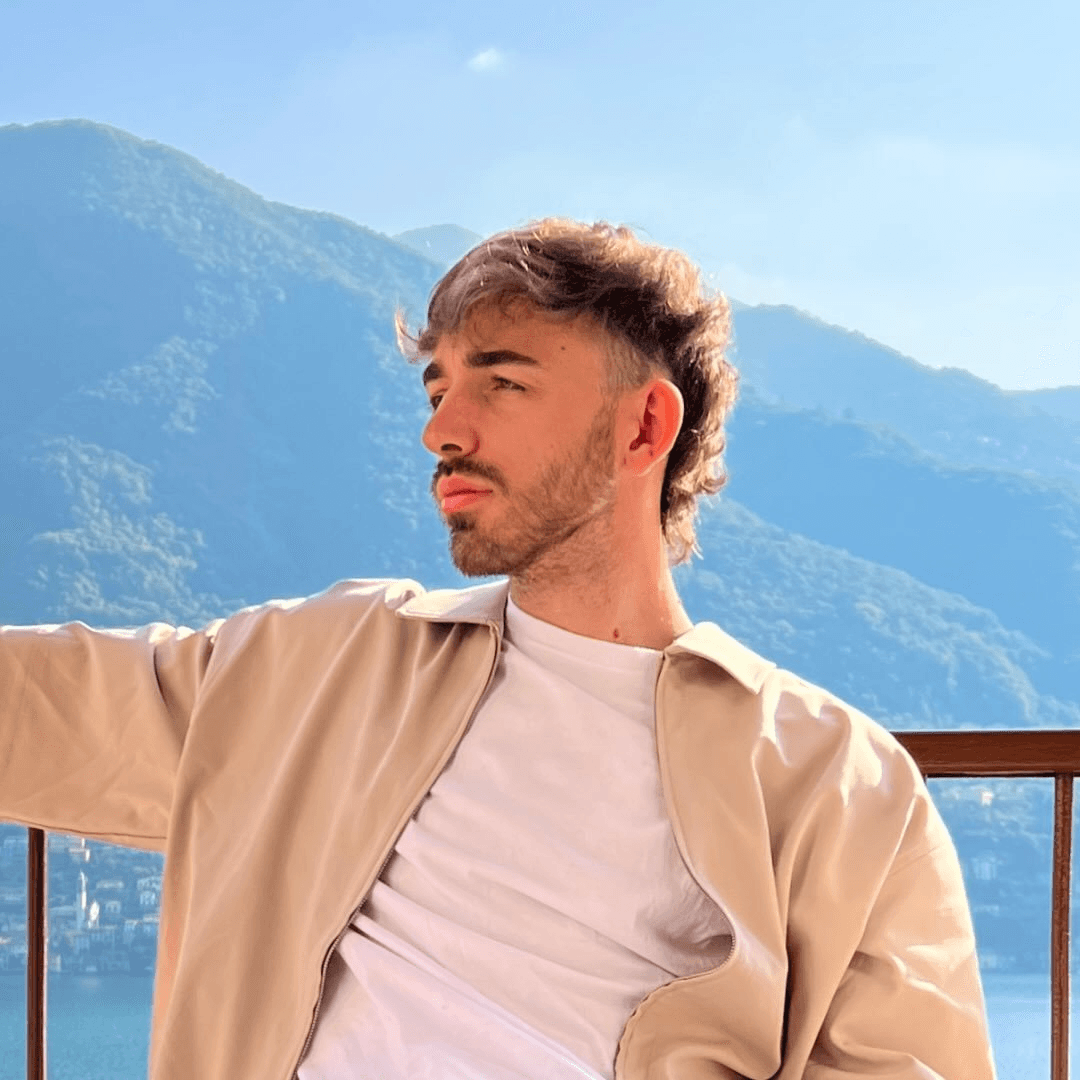 Woman in sunglasses poses against vibrant red background, wearing a beige blazer and black top. Sunlight casts dynamic shadows, creating a stylish, confident mood.