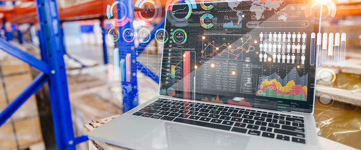 A laptop displaying data on a table in a brightly lit warehouse with blue shelving in the background.