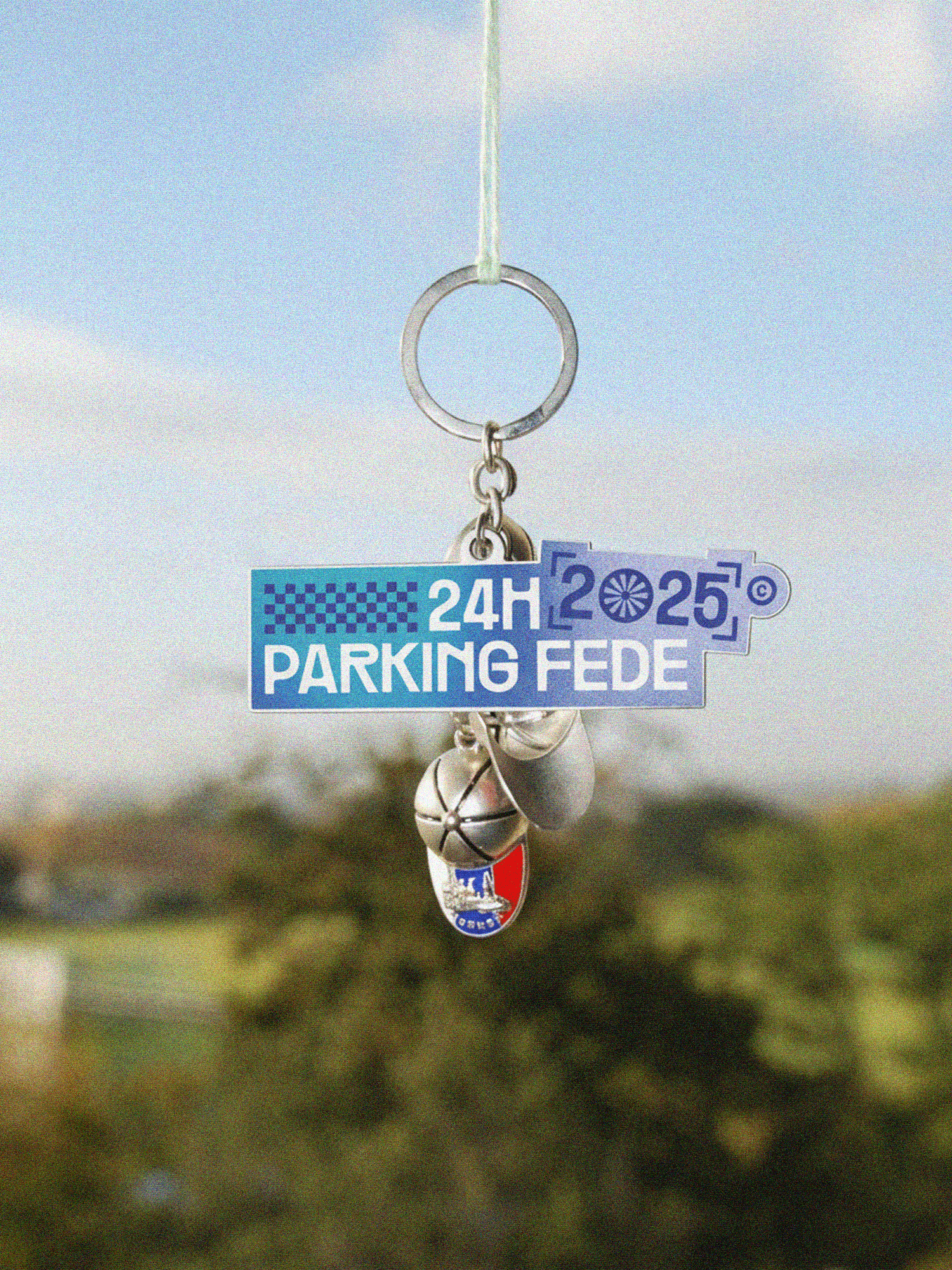 Un gros plan sur un porte-clés suspendu par un fin cordon, sur fond extérieur flou et doux. Le porte-clés en émail arborant le logo des 24H Parking Fédé 2025 est placé au centre de la composition, accompagné de quelques breloques métalliques qui apportent du poids visuel et renforcent le réalisme de la scène.