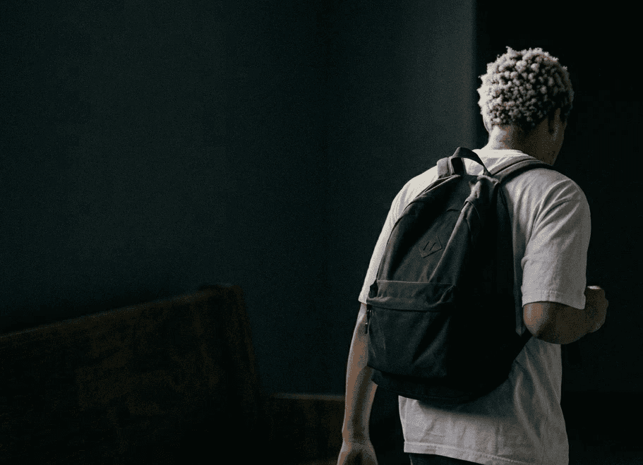 A person with curly hair walks down a dimly lit hallway wearing a casual t-shirt and a backpack, creating a mysterious and introspective atmosphere.