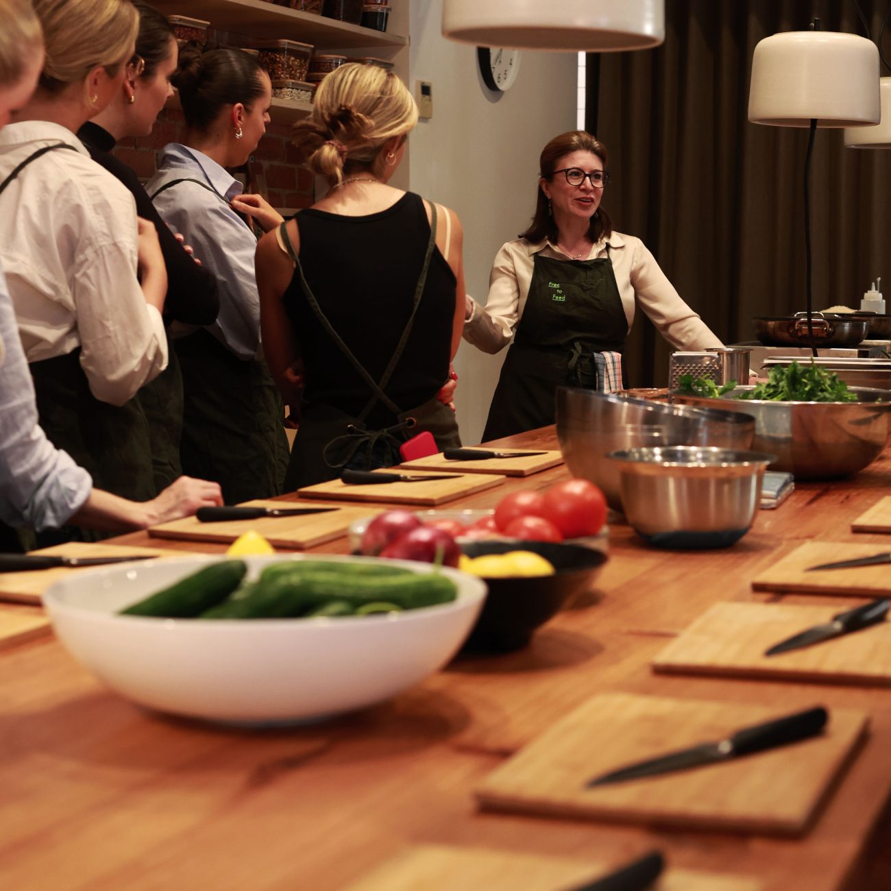 Chef with a "Free to Feed" apron speaking to a group gathered around a kitchen bench set with chopping boards, vegetables and knives