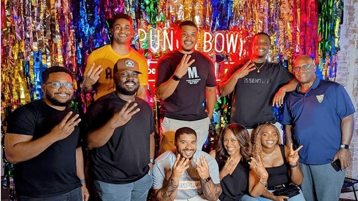 A group of people smiling and posing in front of a colorful backdrop with multicolored foil streamers and a neon sign that reads "PUNCH BOWL," creating a festive and vibrant atmosphere.