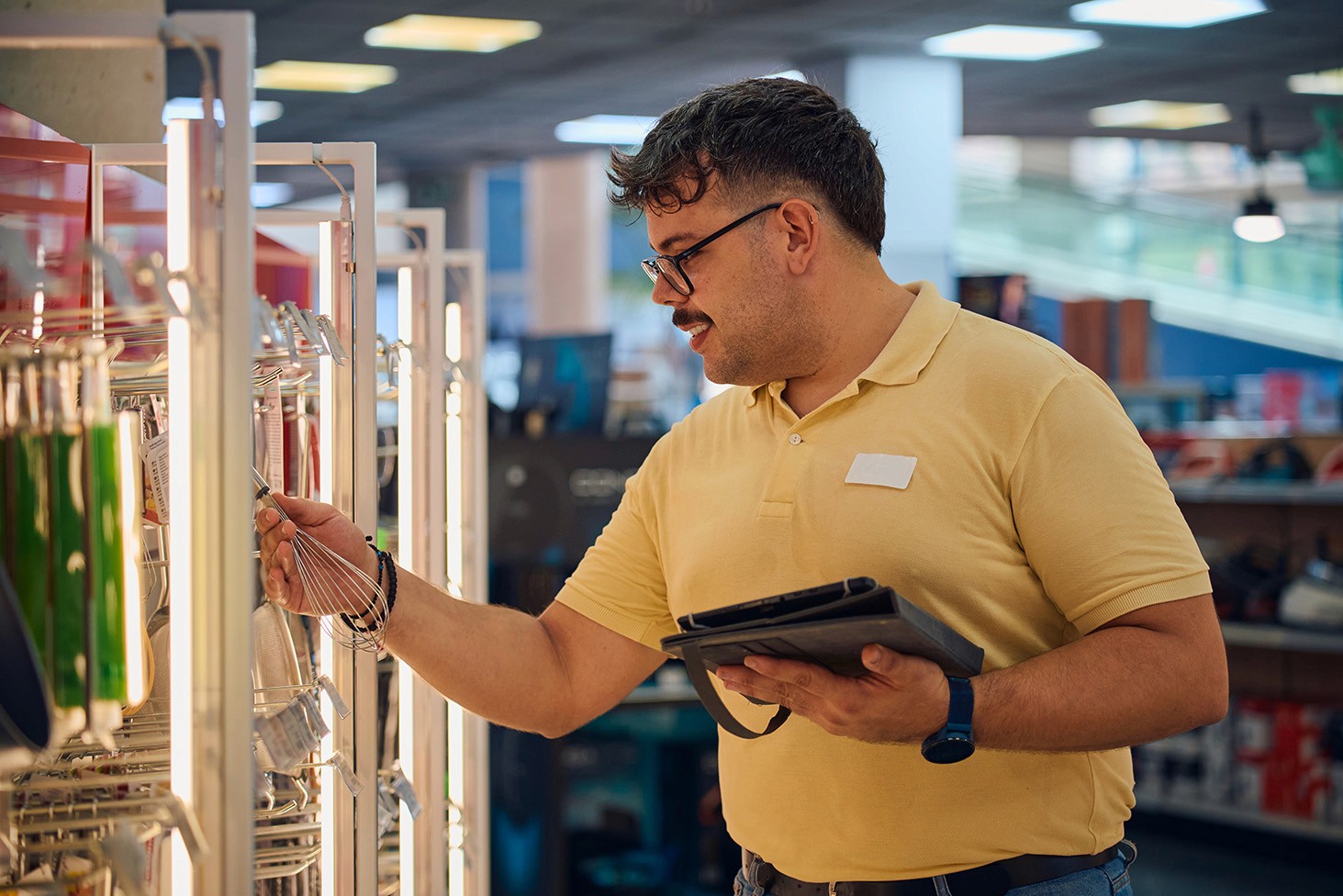 A store employee with glasses and a mustache examines a product on the shelf while holding a tablet.