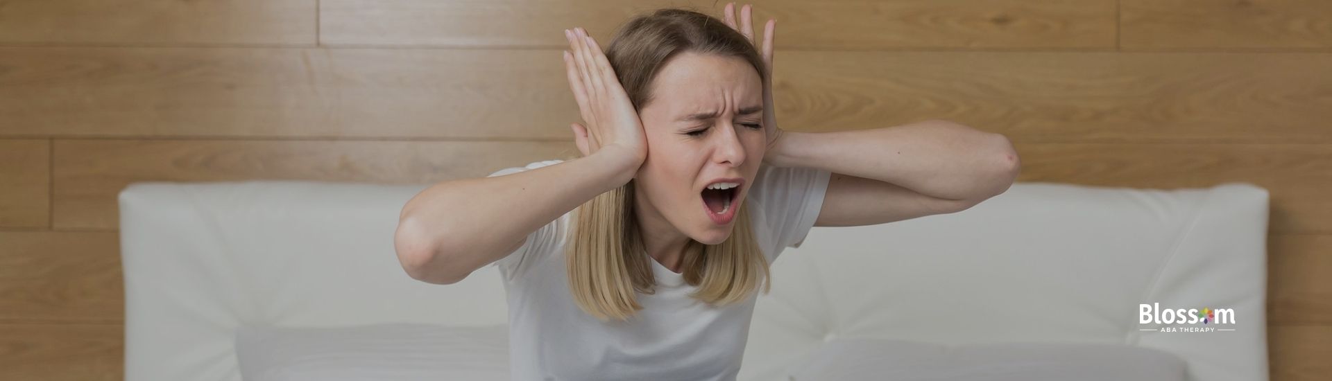 A woman sitting cross-legged on a bed covers her ears and yells in frustration.