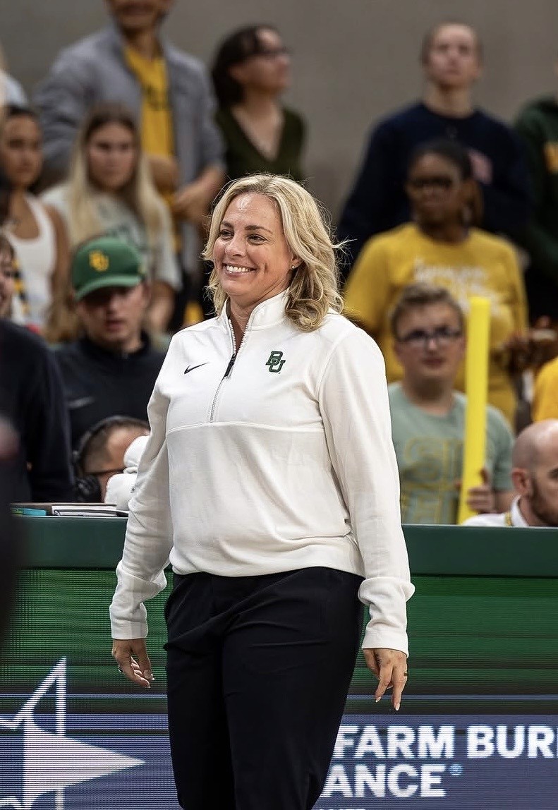 Nicki Collen in a white Baylor University pullover and black pants stands courtside at an indoor sports event. Spectators in yellow and green shirts are visible in the background.