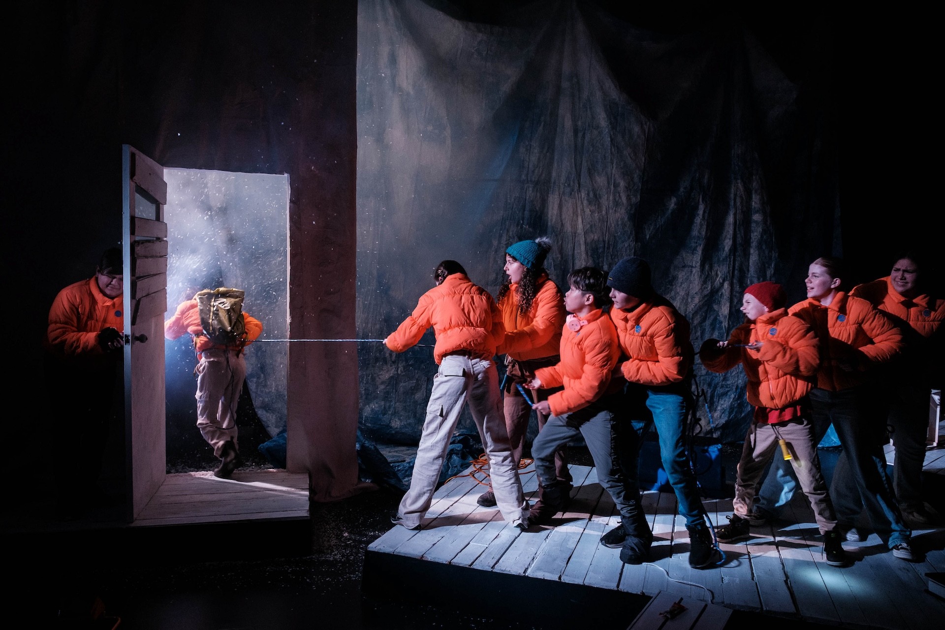 A line of young performers in bright orange puffer jackets brace themselves on a wooden platform, pulling tightly on a safety rope that leads through an open doorway into a bright, snowy blizzard where another performer is stepping outside.