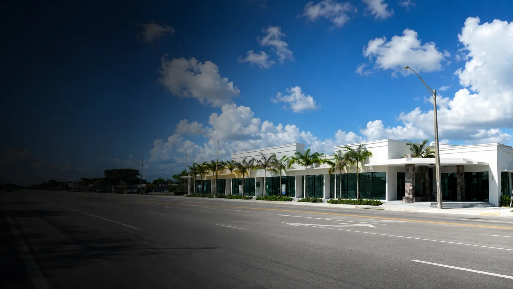 White single-story commercial retail plaza with glass storefronts and palm trees lining an empty Miami street under blue sky