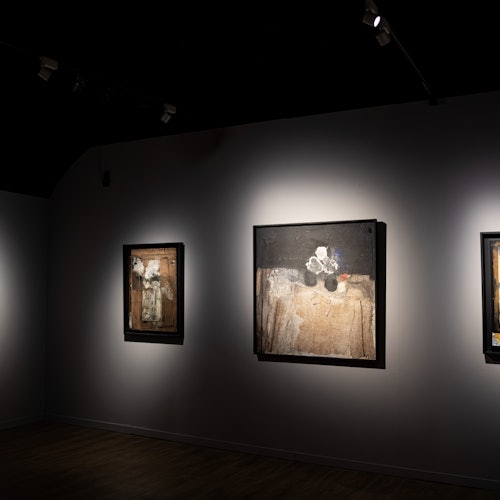 Gallery ng sining na may tatlong abstract na mga pagpipinta na naka-frame at may ilaw sa isang madilim na kulay-abong pader. Ang espasyo ay may sahig na gawa sa kahoy at malabong ilaw.