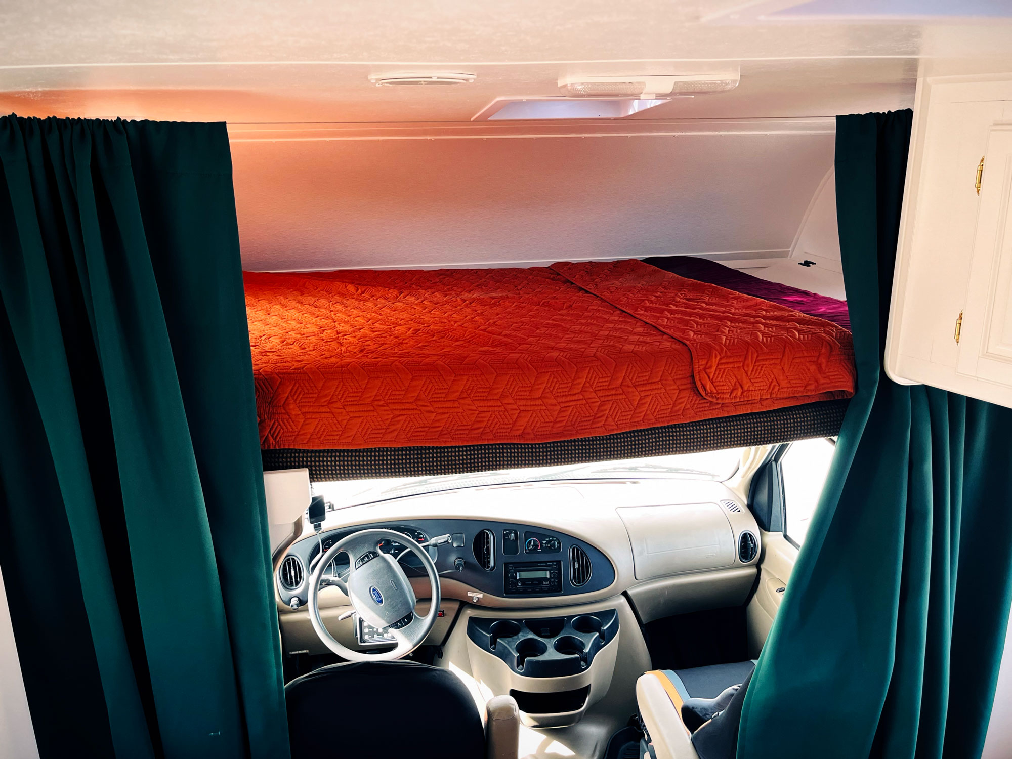 RV interior showing a front driving area with a beige dashboard and steering wheel. Above, a red blanket covers an overhead bunk bed.