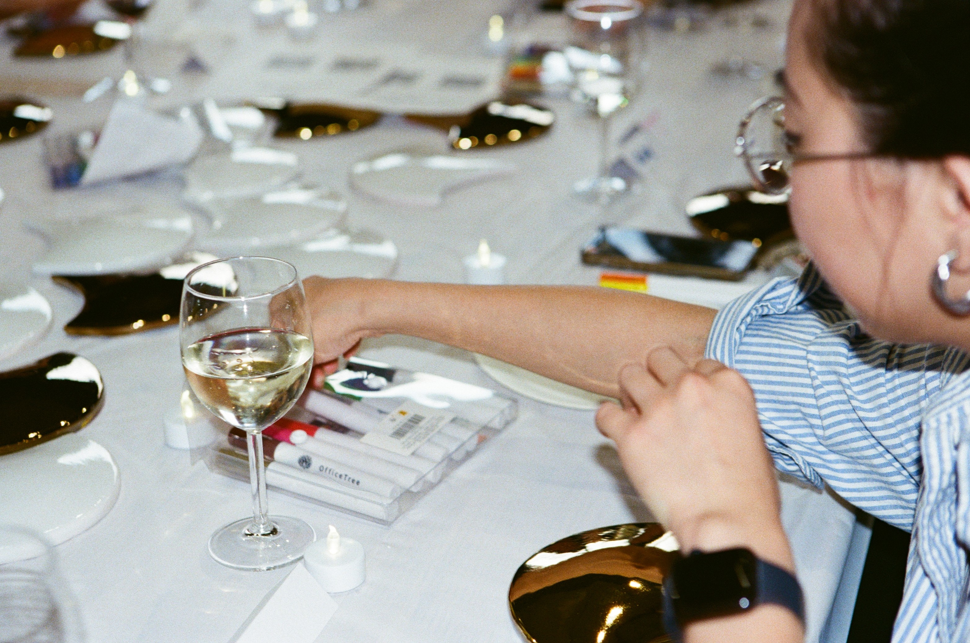 A guest in a blue striped shirt reaching across the table for a glass of white wine, blank gold and white fan tiles, a marker set and tealights laid out around her.