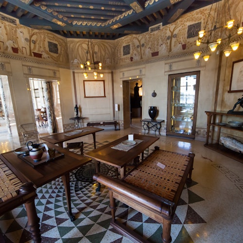 Ornate room with wooden furniture, geometric tiled floor, frescoed walls, chandeliers, and an open door leading to a bright area.