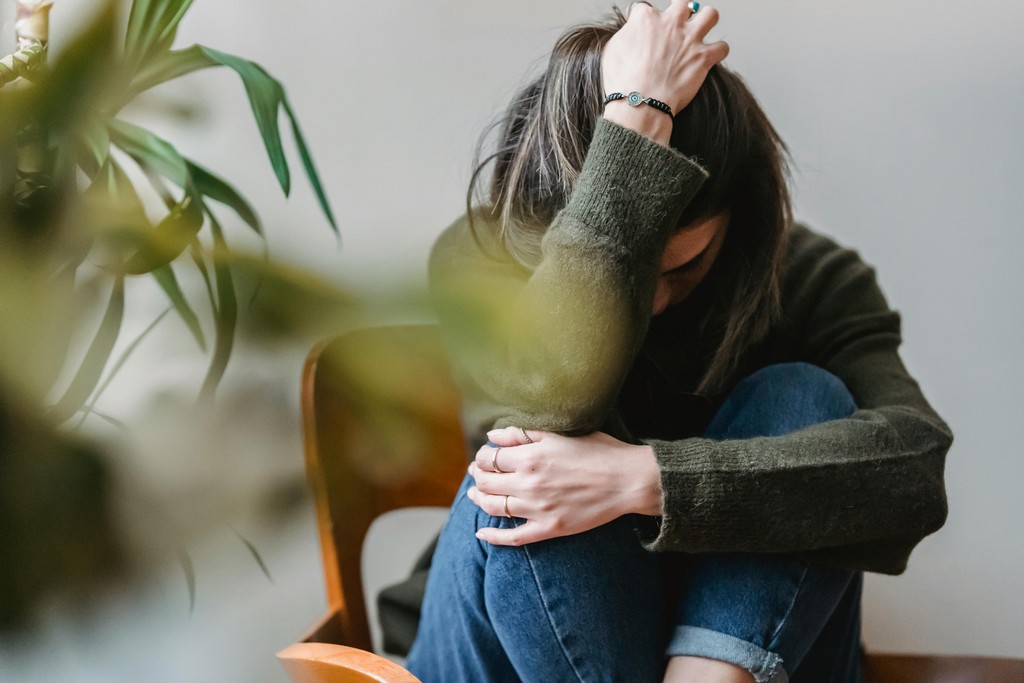 A person sitting with their head down, looking distressed, next to a plant in a softly lit room.