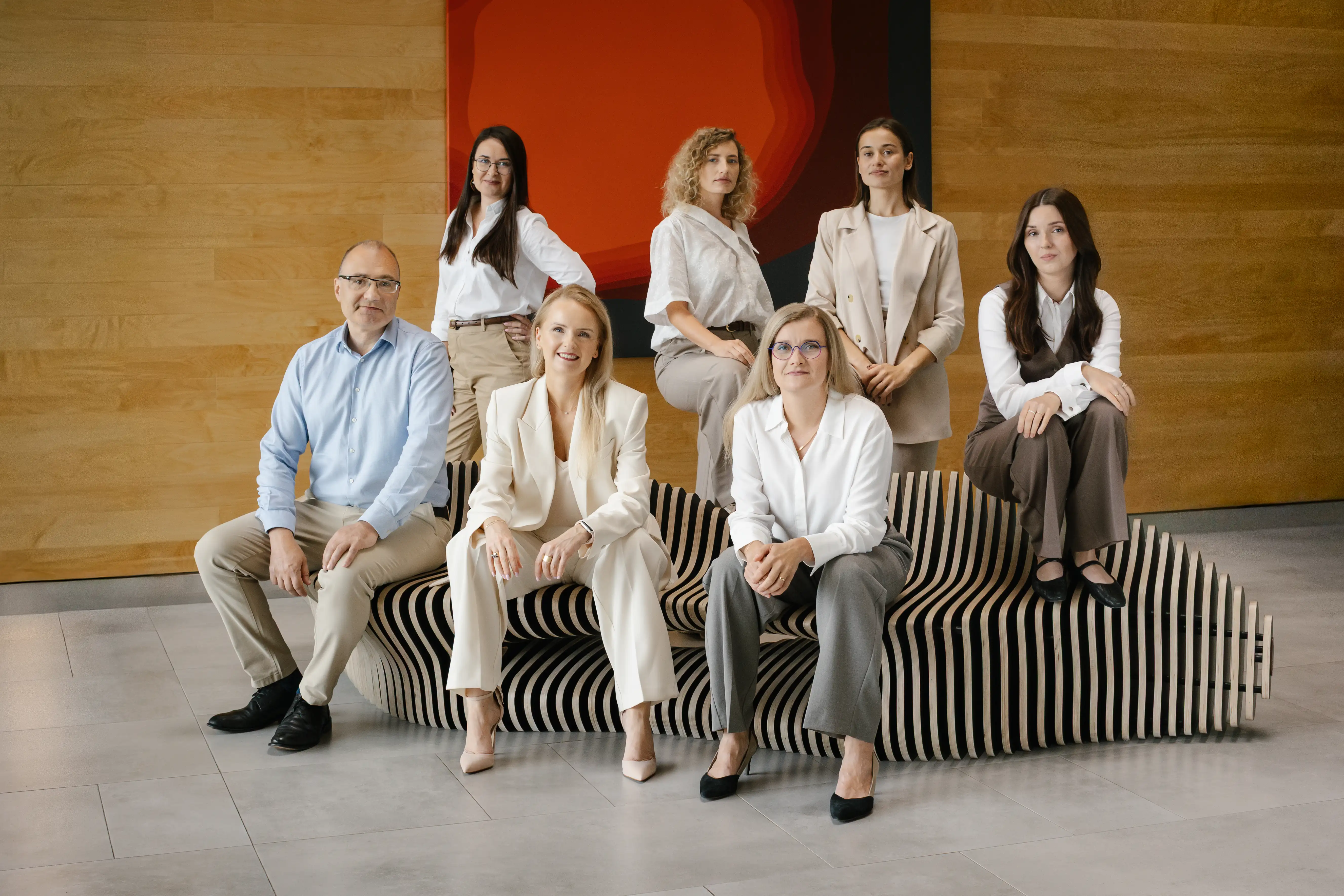 Siedem profesjonalnie ubranych osób pozuje razem w pomieszczeniu na i wokół nowoczesnej, falistej ławki, z drewnianą ścianą i dużą abstrakcyjną czerwoną grafiką w tle.