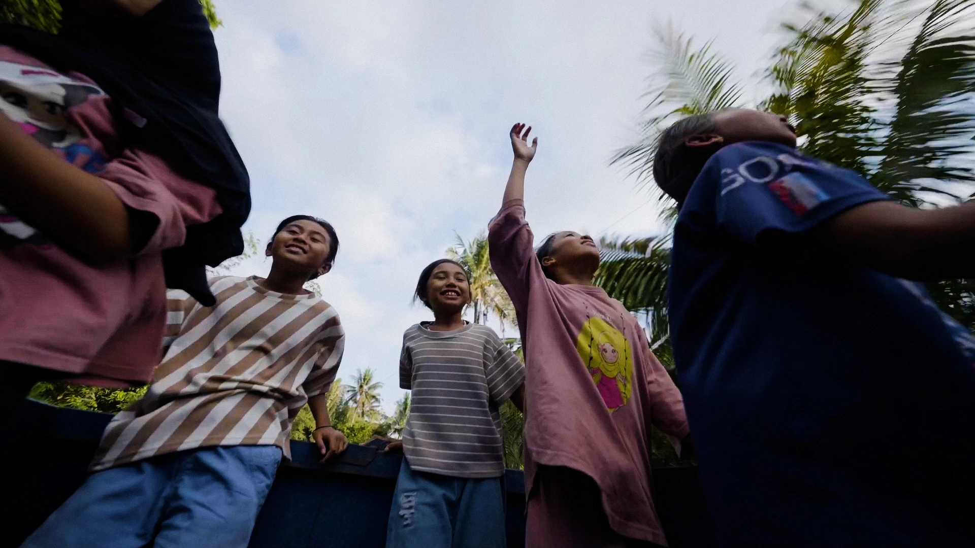 Children laughing outdoors, wearing colorful shirts, with palm trees and a blue sky in the background, capturing a carefree, joyful moment.