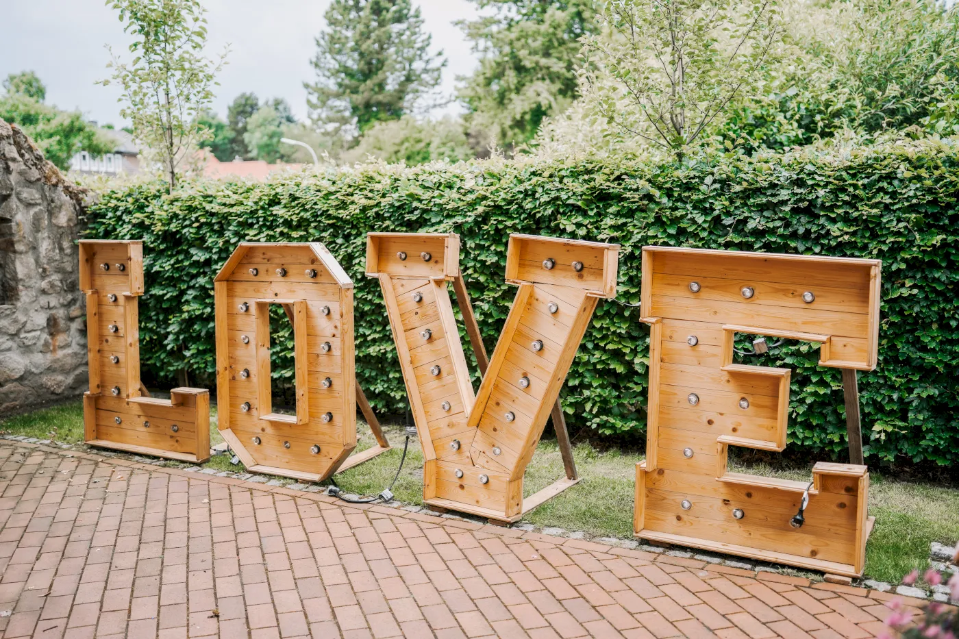 Große Holzbuchstaben bilden das Wort "LOVE" mit Lichtern, die vor einer Hecke und einem Ziegelboden stehen.